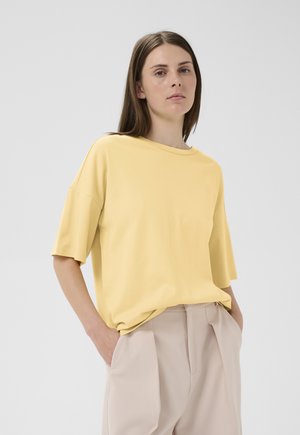 Noorpruunide sirgete juustega noor naine kannab lahtist kollast lühikeste varrukatega toppi ja beeže kõrge vöökohaga plisseeritud pükse, käed taskutes.