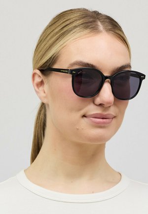 Blonde Frau mit zurückgebundenem Haar, die schwarze quadratische Sonnenbrille und ein weißes T-Shirt mit Rundhalsausschnitt trägt, neutraler Hintergrund.