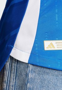 Sininen ja valkoinen urheiluasu, jossa on strukturoitu muotoilu, teksturoitu kangas ja kultainen brodeerattu brändilogo valkoisella kentällä.