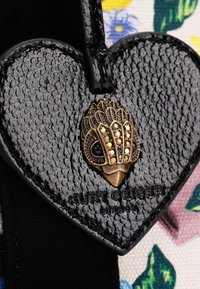 Etichetta di pelle nera a forma di cuore con una finitura testurizzata e un accento decorativo color oro con dettagli intricati e un occhio di pietra preziosa. Marchio visibile.