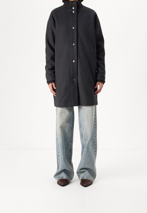 Personne portant un long manteau gris foncé avec des boutons-pression, un jean large bleu clair et des chaussures pointues marron, debout devant un fond blanc.