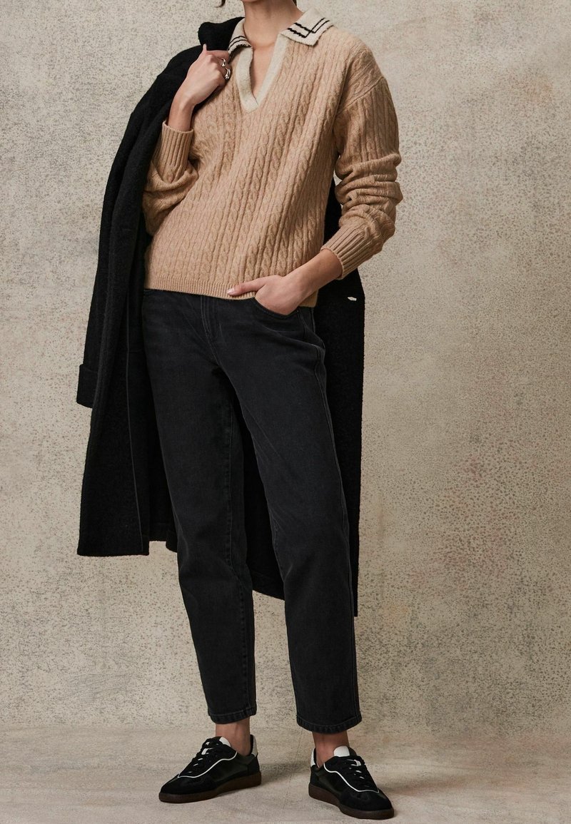 Personne portant un pull beige en maille torsadée avec un col blanc, un jean noir fuselé, des baskets noires, tenant un manteau noir sur une épaule.