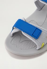 Pelēki sandaļi ar teksturētu zoli, ar ziliem regulējamiem siksnām un dzeltenu akcentu. Izgatavoti no viegla materiāla, lai nodrošinātu komfortu.