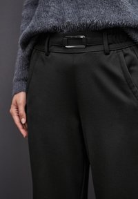 Schwarze Hose mit einer eleganten Textur, ausgestattet mit einem glatten Gürtel und einer metallischen Schnalle. Weicher, bequemer Stoff mit seitlichen Taschen.