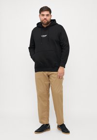 Zwarte katoenen hoodie met logo op de borst, kangoeroezak, capuchon met trekkoord, gecombineerd met tan broeken en zwarte sneakers. Effen achtergrond.