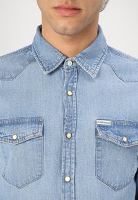 Camisa de mezclilla azul con cuello abotonado, dos bolsillos en el pecho y botones a presión dorados. Cuenta con el logo "GUESS JEANS" en el bolsillo.