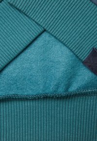 Textil verde azulado con textura de canalé en la parte superior y tejido suave y afelpado debajo. Etiqueta de tela azul oscuro con un patrón sutil en el lateral.