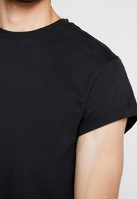Nærbillede af en person iført en enkel sort t-shirt med korte ærmer og rund hals mod en neutral baggrund.