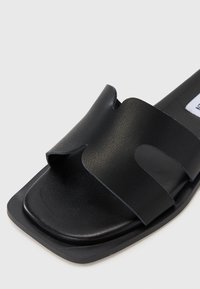 Fekete lapos szandál, négyzet alakú orral, textúrált felső pánttal és sima talppal. Minimalista dizájn, látható fémalkatrészek nélkül.