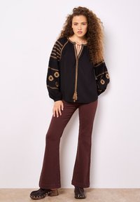 Blusa nera con ricami dorati e nappine, dotata di lunghe maniche a sbuffo. Abbinata a pantaloni svasati bordeaux e zoccoli neri.