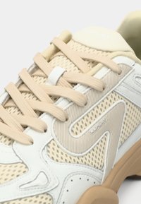 Beige athletic shoe with mesh and rubber overlay, featuring a textured design, flat laces, and a white logo on the tongue.