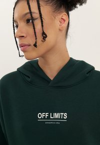 Green hoodie made of soft fabric, featuring a drawstring hood and printed text "OFF LIMITS DANGEROUS AREA" in white on the front.