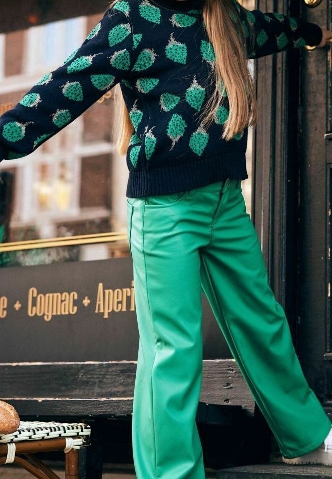 Personne portant un pull noir avec un motif de houblon vert et un pantalon vert vif, debout les bras tendus près de l'entrée d'un café.