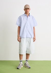Lys blå kortermet skjorte med knapper, avslappet passform, sammen med lyse shorts. Grå sneakers med hvite og svarte stripete sokker, beige caps.