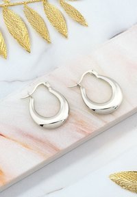 Silvriga hoops örhängen med en slät, reflekterande yta och en böjd design, visade upp på en marmorliknande yta med guldinslag.