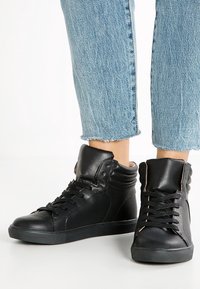 Černé kožené vysoké tenisky mají zaoblenou špičku, šněrování a texturované detaily. Silná gumová podešev zajišťuje přilnavost a odolnost.