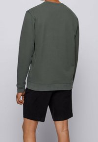 Grön sweatshirt med långa ärmar, ribbad rund halsringning och rak nederkant; bärs med svarta shorts. Slät tyg med synliga sömnadsdetaljer.