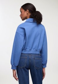 Felpa blu corta realizzata in tessuto morbido con colletto alto, vestibilità rilassata e orlo a costine. Abbinata a jeans di denim scuri con tasche posteriori.