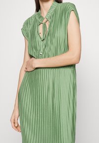 Grünes, plissiertes Kleid mit hohem Kragen, kurzen Ärmeln und einer Vorderseite mit Schlüsselloch-Bindedetail. Glatte Textur mit fließender Form.