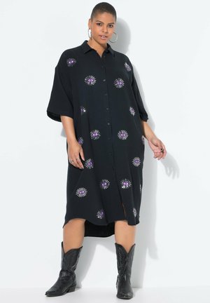 Schwarzes Hemdkleid mit kurzen Ärmeln, das lila Augenmuster und einen strukturierten Stoff aufweist. Kombiniert mit schwarzen Cowboy-Stiefeln.