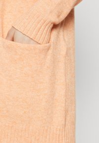 Ruka zasunutá v kapse měkkého pleteného svetru v barvě broskve s žebrovanými manžetami a lemem.