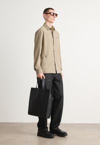 Camicia button-up beige con maniche lunghe, abbinata a pantaloni in denim scuri. Una borsa tote in pelle nera è tenuta in una mano. Occhiali da sole marroni completano il look.