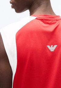 Κοντινό πλάνο ενός ατόμου που φοράει αμάνικο κόκκινο και λευκό πουκάμισο με ένα μικρό λευκό λογότυπο στο πάνω μέρος της πλάτης.