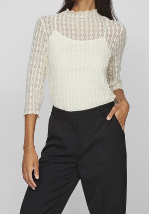 Femme portant un haut en dentelle blanche transparente à manches 3/4 superposé à un caraco blanc, associé à un pantalon noir taille haute.