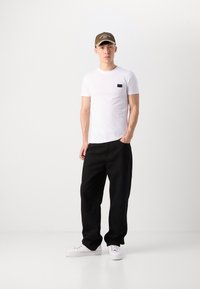 T-shirt bianco a maniche corte con etichetta nera, abbinato a pantaloni neri larghi e scarpe da ginnastica bianche. Un berretto marrone completa l'outfit.