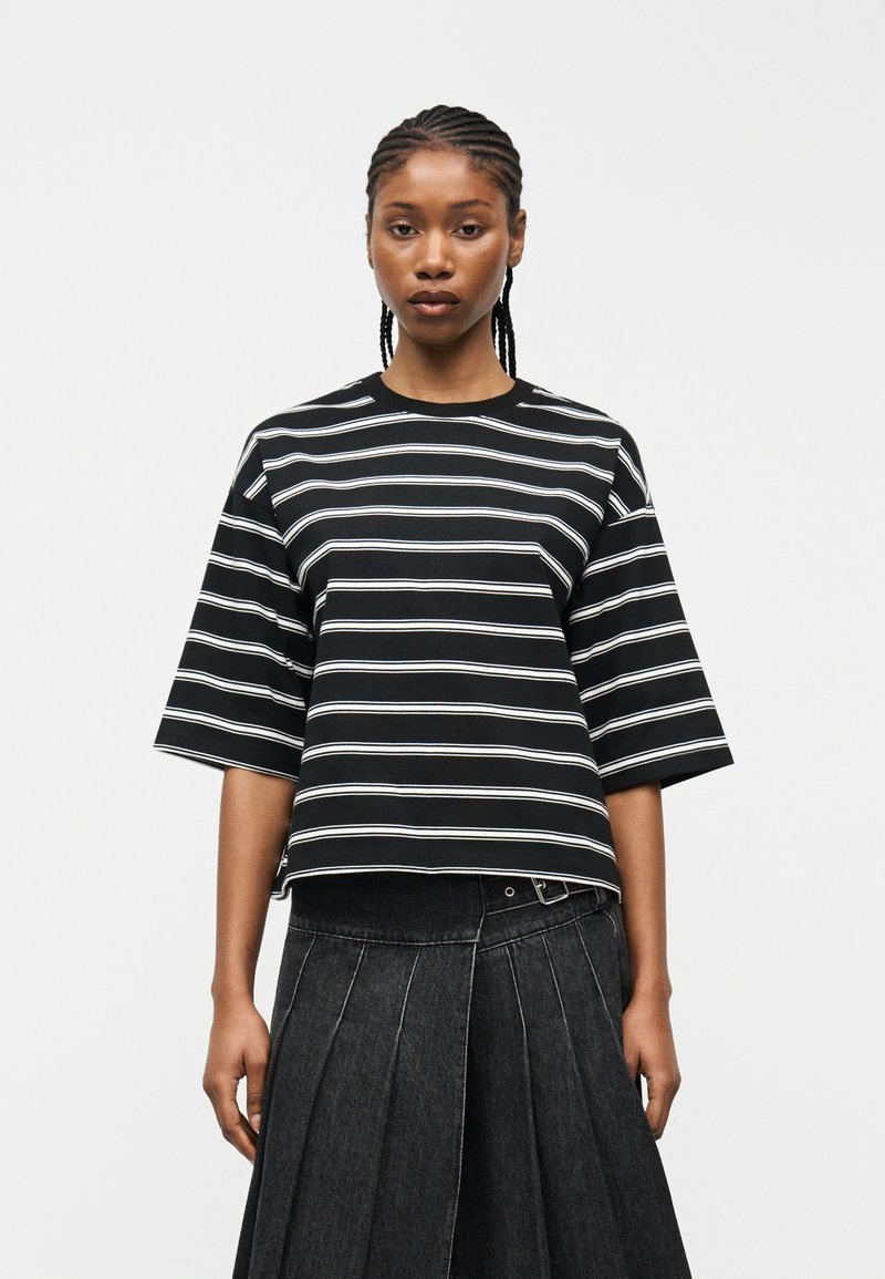 Γυναίκα με πλεξούδες, που φοράει κοντομάνικο ριγέ πουκάμισο σε μαύρο και άσπρο και σκουρόχρωμη πλισέ φούστα με ψηλή μέση, μπροστά σε απλό φόντο.