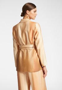 Blazer in satin con sfumature degradé di pesco e crema, caratterizzato da una forma aderente, una cintura in vita e un accento decorativo sul retro.