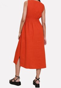 Robe longue orange sans manches avec une taille froncée, des fentes latérales et une texture de tissu lisse. Associée à des sandales à plateforme noires à fines brides.