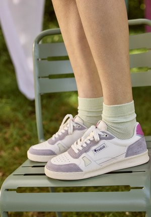 Persoon draagt witte en grijze Faguo sneakers met roze details en crèmekleurige sokken, staand op een lichtgroene metalen buitenstoel.