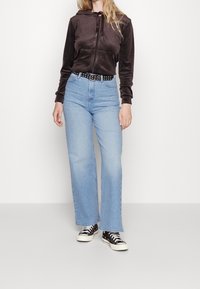 Tmavě hnědá mikina na zip s přední kapsou, kombinovaná se světle modrými rozšířenými džíny a černými teniskami s bílou podrážkou a tečkovaným vzorem.