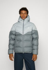 Casaco puffer cinza com um design de dois tons; leve, com textura acolchoada; possui capuz e fecho frontal com zíper; pequeno logótipo no peito.