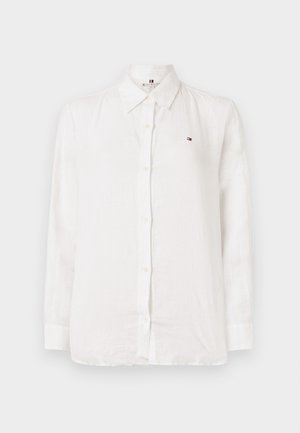 Balta ilgomis rankovėmis marškiniai su sagomis, apykakle, mažas siuvinėtas logotipas kairėje krūtinės pusėje ir matomos sagos priekyje.