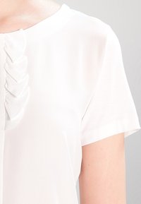 Blusa bianca a maniche corte con scollatura rotonda e dettagli in volant verticali sul davanti, realizzata in un tessuto morbido e leggero.