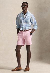 Homme portant des lunettes de soleil, chemise à carreaux bleue avec un pull bleu clair posé sur les épaules, short rose, ceinture marron, montre et mocassins marron debout.