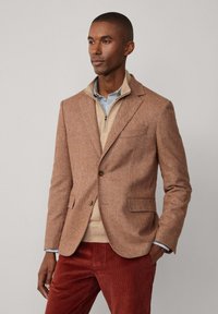 Blazer in tweed marrone con rever a punta, chiusura con un solo bottone e tre tasche frontali, abbinato a un maglione crema e pantaloni in velluto a coste rossi.
