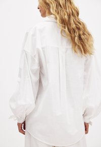 Weißes, locker sitzendes Blusenshirt aus leichtem Stoff, mit einem Rückendrapierung, Ballonärmel und Bindebändern an den Manschetten.