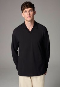 Svart långärmad polo med V-hals. Tyget ser slätt ut, med ribbad krage. Modellen bär ljusa byxor och står mot en neutral bakgrund.