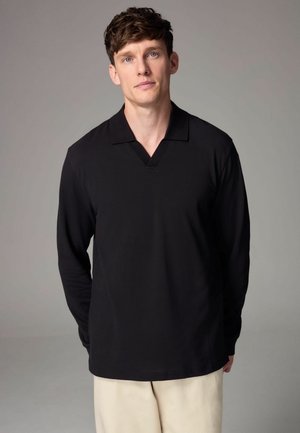 Schwarz langärmeliges Poloshirt mit V-Ausschnitt. Der Stoff wirkt glatt, mit geripptem Kragen. Das Modell trägt hellfarbene Hosen und steht vor einem neutralen Hintergrund.