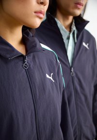 Svart atletiska jackor med dragkedjor framtill, prydda med vita Puma-loggor och turkosa detaljer. Mjuk textur och avslappnad passform, bärs av två personer.