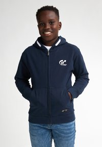 Felpa con zip color navy, tasche frontali, cappuccio con cordino e dettagli logo bianchi. Indossata con jeans di denim azzurro chiaro.