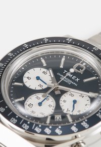 Chronograph watch with a stainless steel band, blue dial, white subdials, silver hands, and a black bezel featuring minute markings.