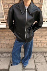 Giubbotto in finta pelle nera con grande colletto e zip frontale, abbinato a jeans blu larghi e scarpe nere a punta.
