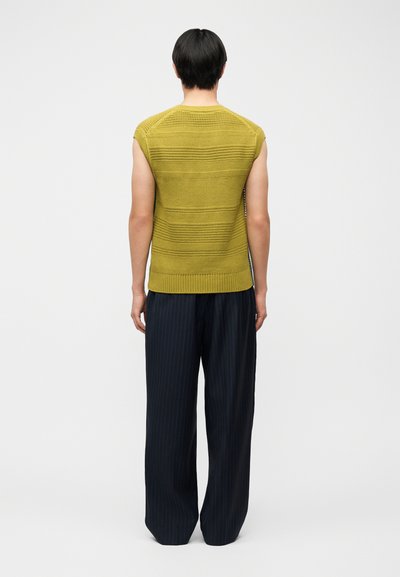 Personne portant un haut en tricot moutarde sans manches et un pantalon large à fines rayures noires, debout de dos devant un fond uni.