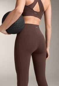 Nő barna sportleggingsben és rövid felsőben, kezében egy texturált fekete edzőlabdával, semleges háttér előtt.