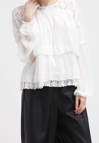 Blouse blanche à couches avec manches en dentelle transparente et bordure en dentelle à l'ourlet. Portée sur un pantalon noir ample. Texture légère et aérienne.