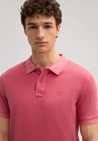 Różowa koszulka polo z kołnierzykiem, trzema guzikami z przodu oraz haftowanym logo "JOOP! Jeans" po lewej stronie klatki piersiowej. Miękka tekstura materiału.